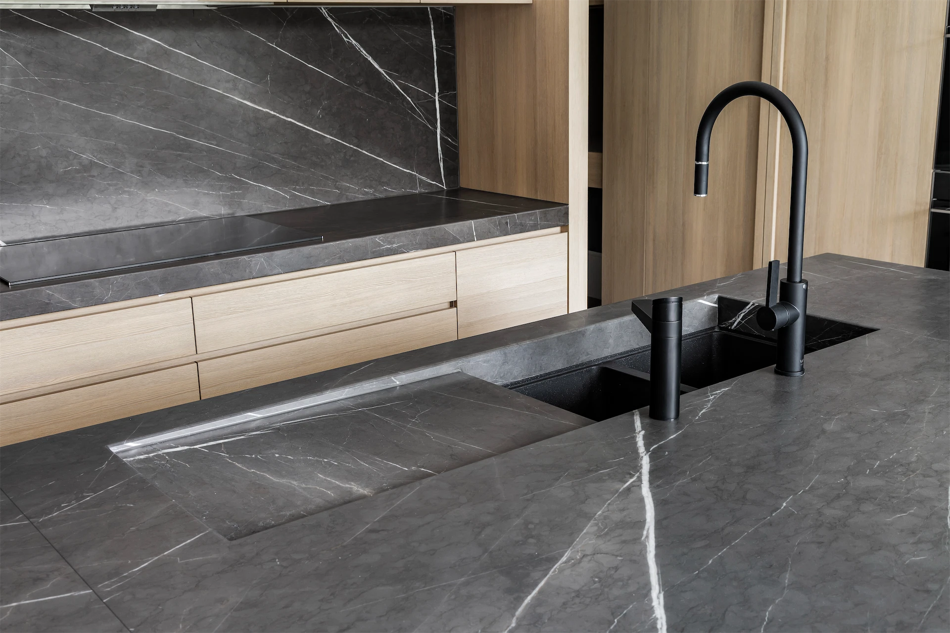 Modern kitchen renovation featuring dark marble countertops, integrated sink, light wood cabinetry, and matte black fixtures